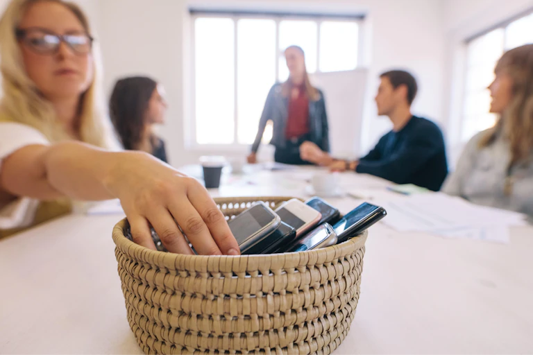 A basket of discarded smartphones with people sitting around a table behind it.