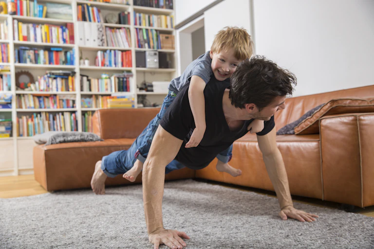 Many with a small child on his back doing a plank in a living room.
