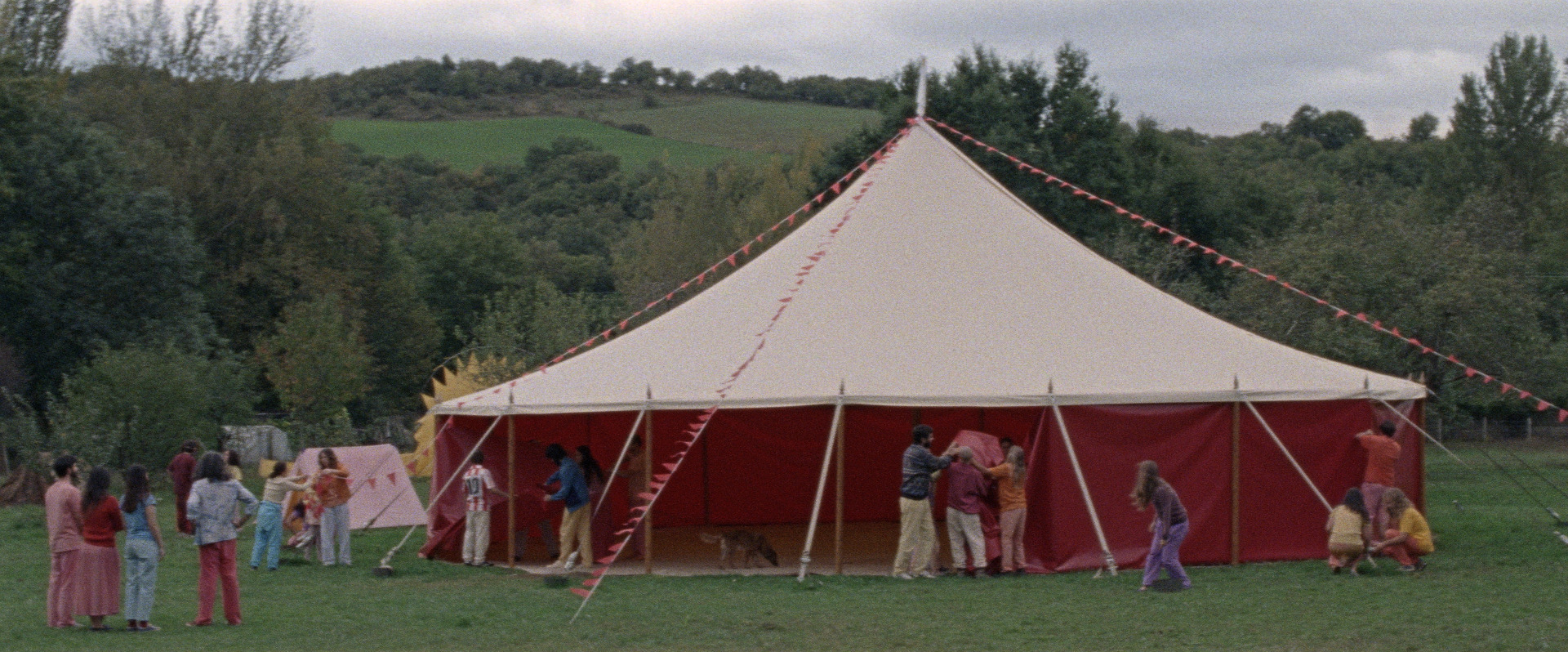 A large beige and red circus tent set up on a green meadow surrounded by a forest, with people working around it. A scene from Aro Beria's film about communes in Spain.