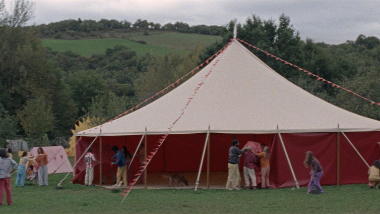 'Aro Berria', a cinematic reminiscence of the communes that puzzled the circle of relatives and posed a utopia 1 A large beige and red circus tent set up on a green meadow surrounded by a forest, with people working around it. A scene from Aro Beria's film about communes in Spain.