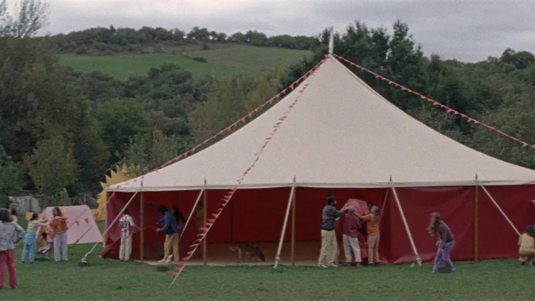 Una gran carpa de circo de color beige y rojo montada en un prado verde rodeado de bosques, con personas trabajando alrededor. Escena de la película Aro Berria sobre comunas en España.