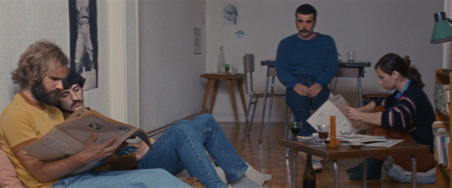 Un grupo de jóvenes con estética años 70 sentados en un salón leyendo un periódico con expresión seria y aburrida en sus rostros. Una escena de la película de Aro Beria.