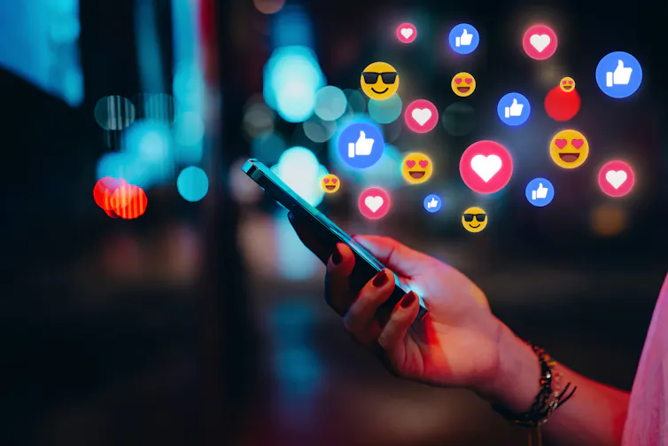 hand holds phone with emojis, hearts, likes etc rising in a cloud around it