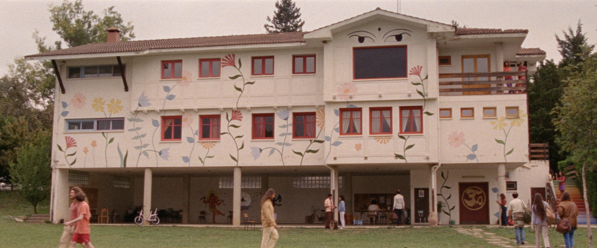 The facade of a large three-story house painted white and decorated with large colorful flowers and two giant eyes on top. Several people are walking on the grass in front of the building.