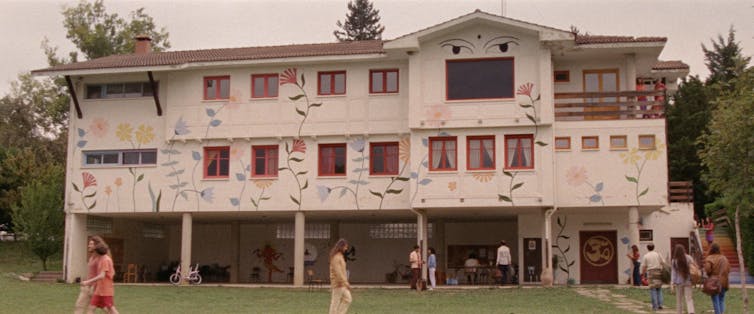 'Aro Berria', a cinematic reminiscence of the communes that puzzled the circle of relatives and posed a utopia 3 The facade of a large three-story house painted white and decorated with large colorful flowers and two giant eyes on top. Several people are walking on the grass in front of the building.