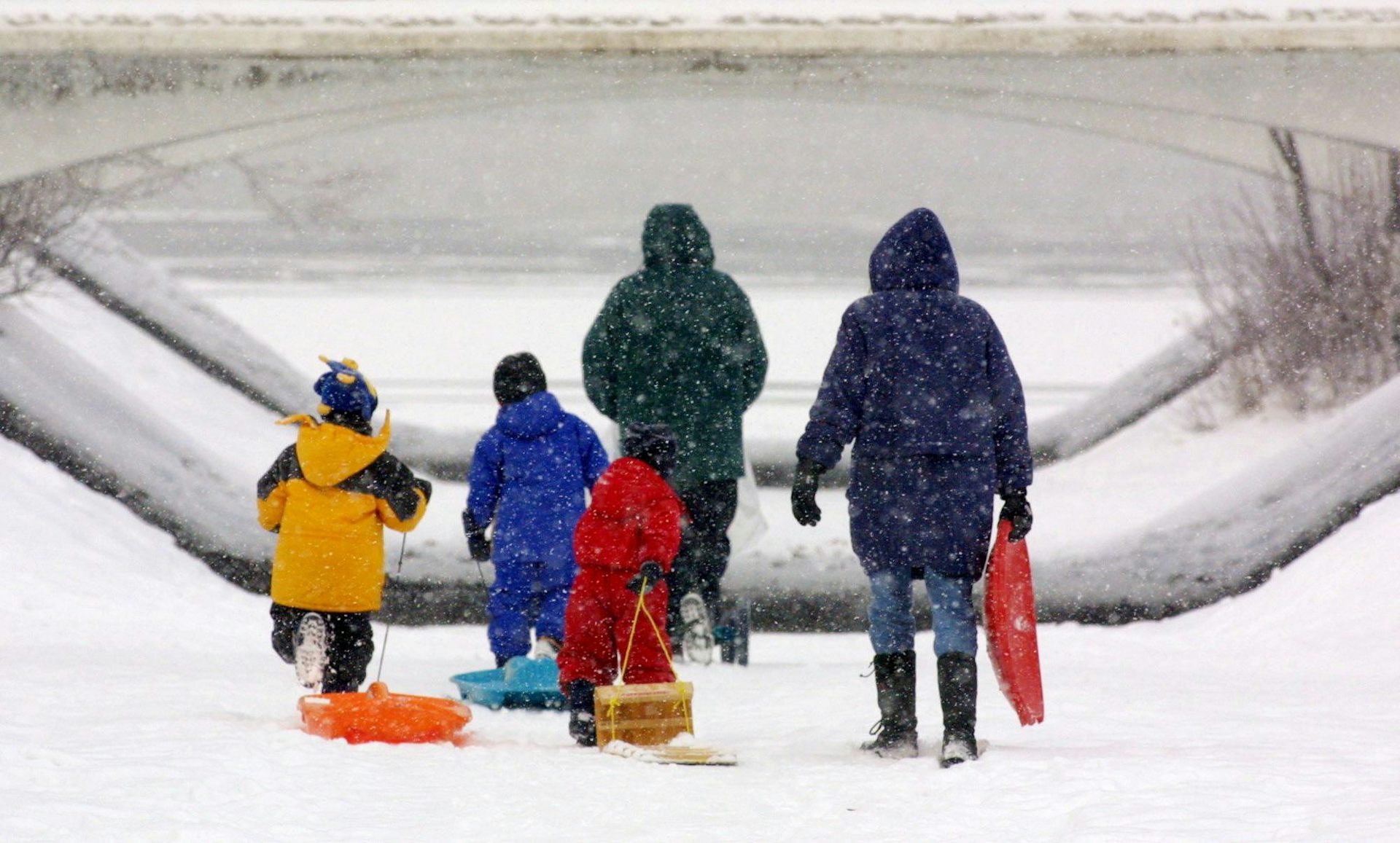 Two adults and three children in winter clothing seen from behind with a toboggan and sliding toys in the snow