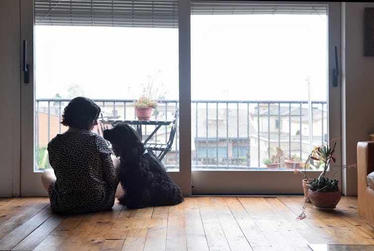 A woman with short brunette hair sits on the floor in front of a sliding door and balcony, as a black dog sits beside her and looks at her.