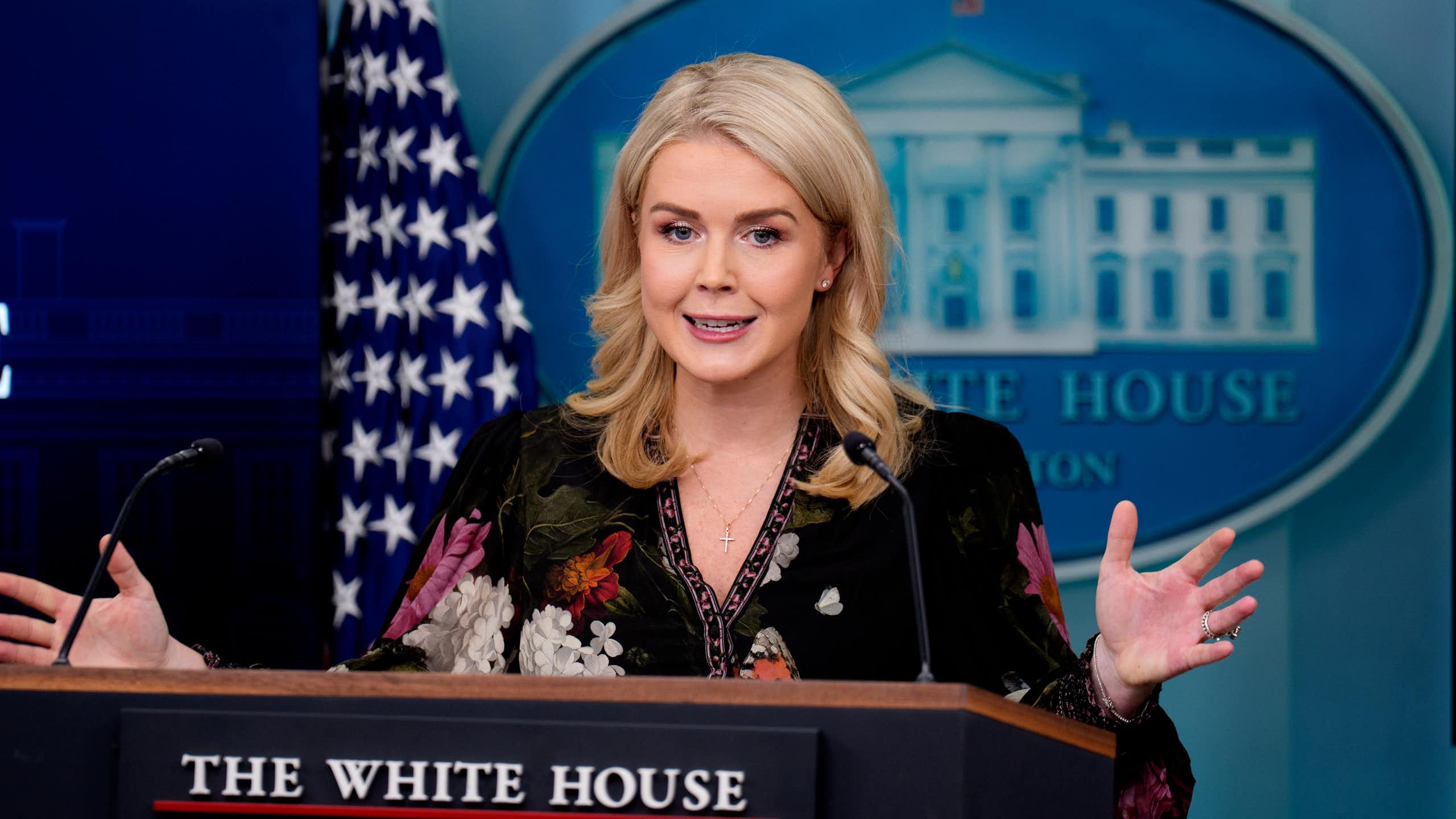 A woman in a dark top with blond hair behind a lectern, with a US flag behind her as well as an image of the White House.