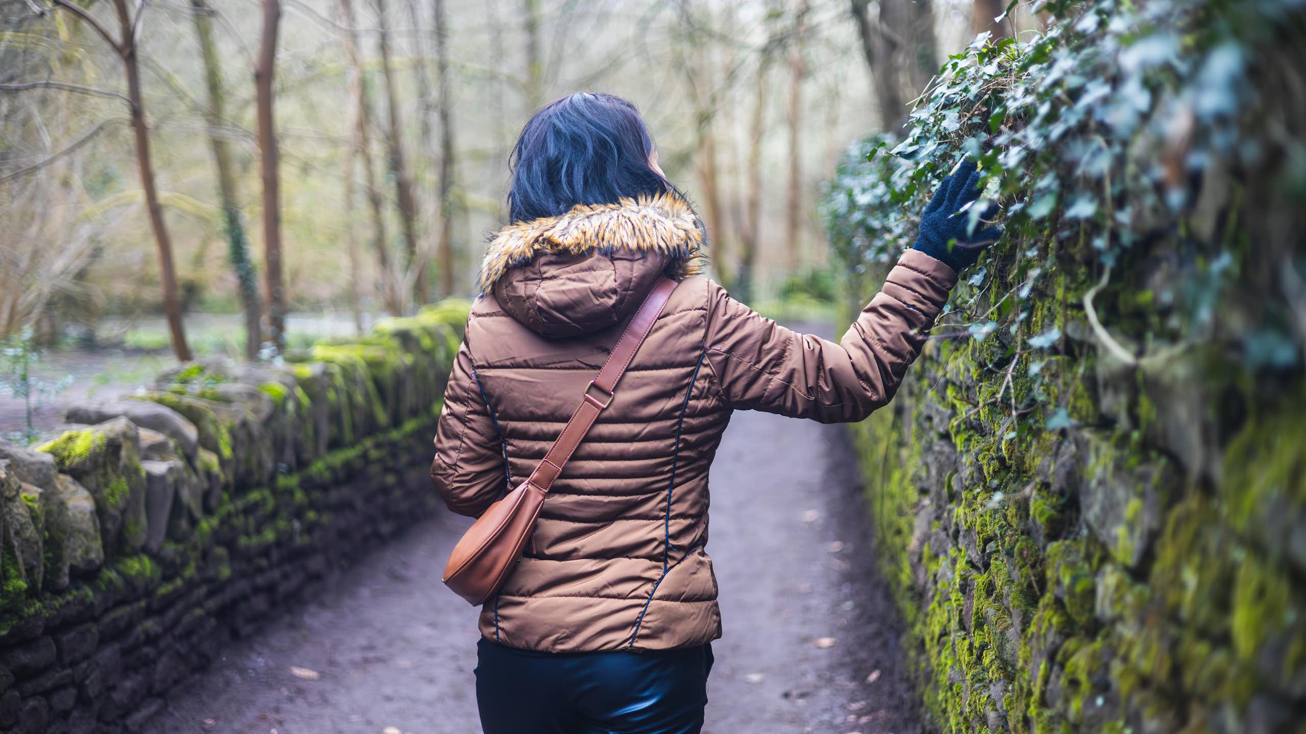 A woman in a winter jacket walks along an ivy-covered wall in a park.
