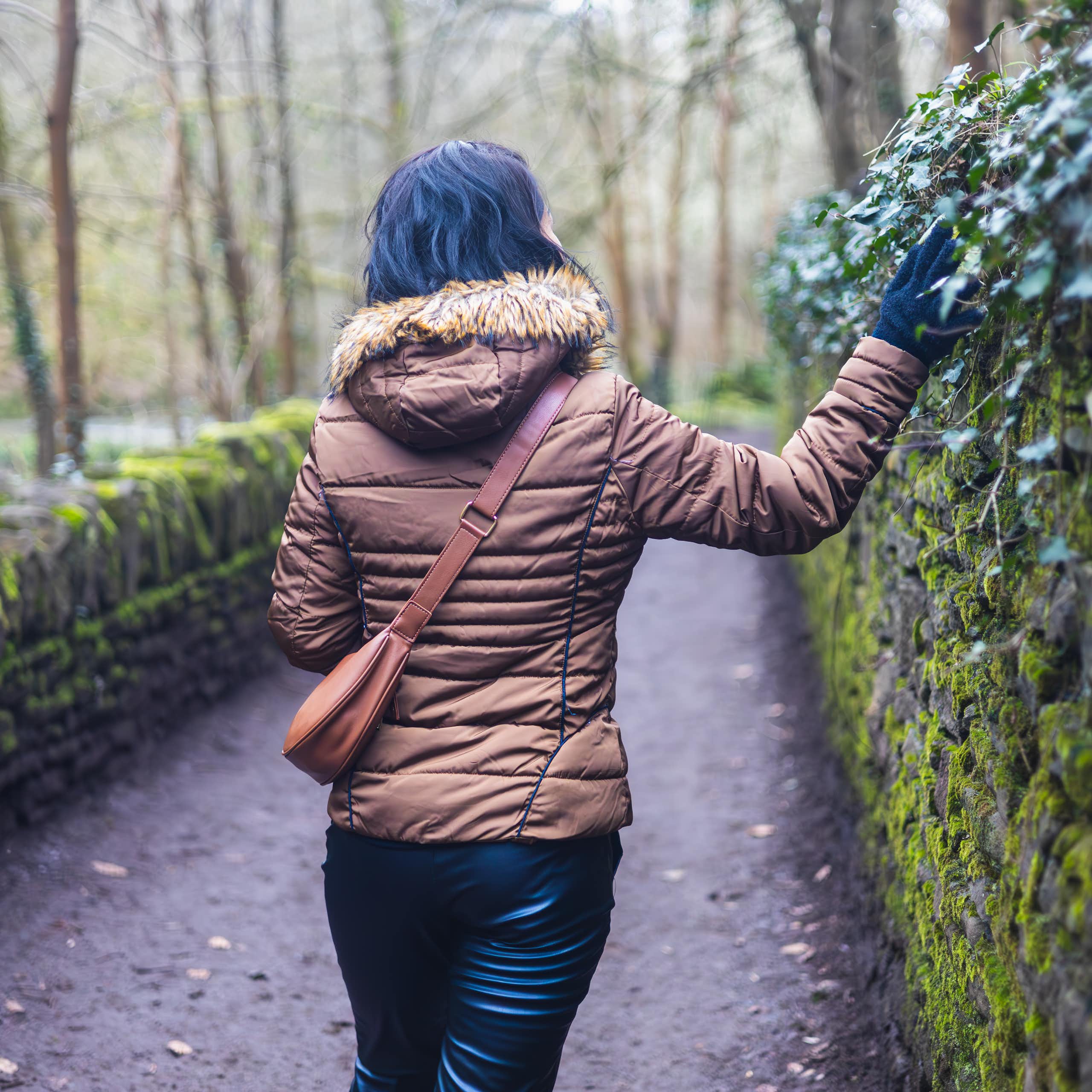 A woman in a winter jacket walks along an ivy-covered wall in a park.