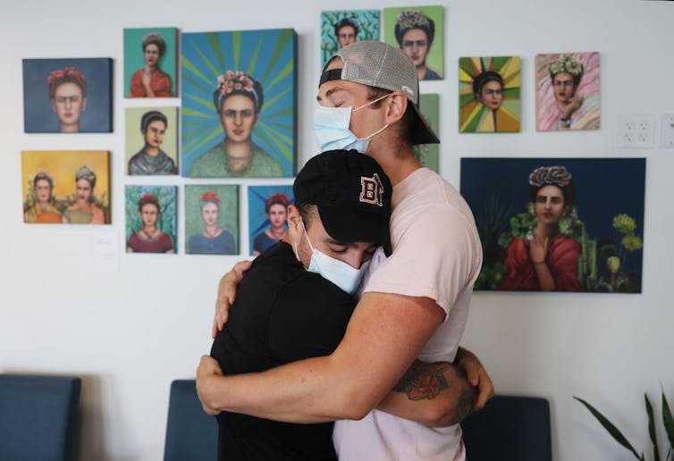 Two people wearing surgical masks embracing, paintings of Frida Kahlo on the wall behind them