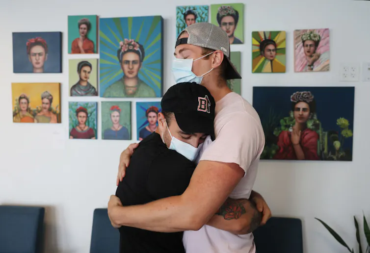 Two people wearing surgical masks embracing, paintings of Frida Kahlo on the wall behind them