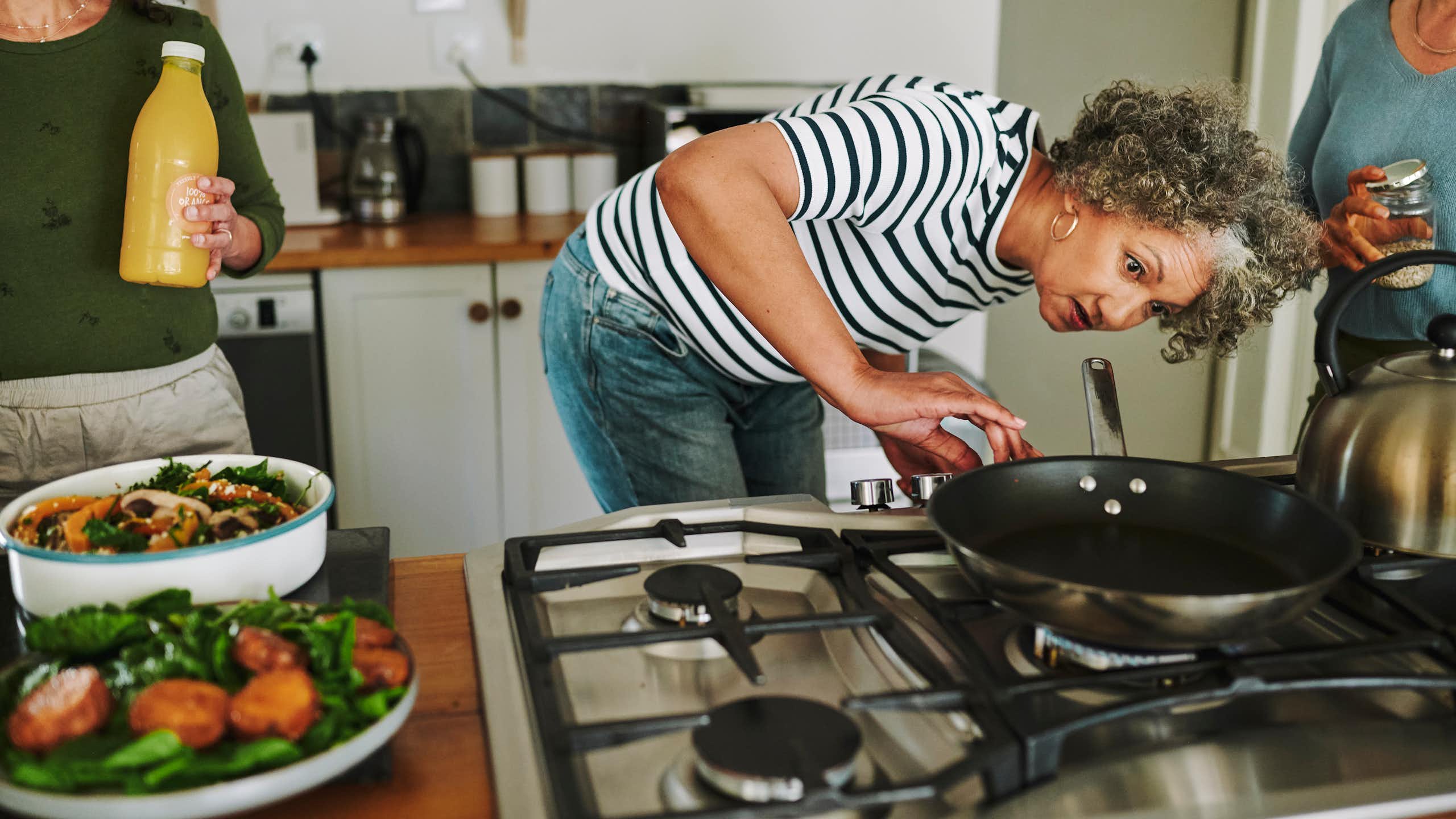 A woman with a black and white striped shirt looks under a pan on a stove top.