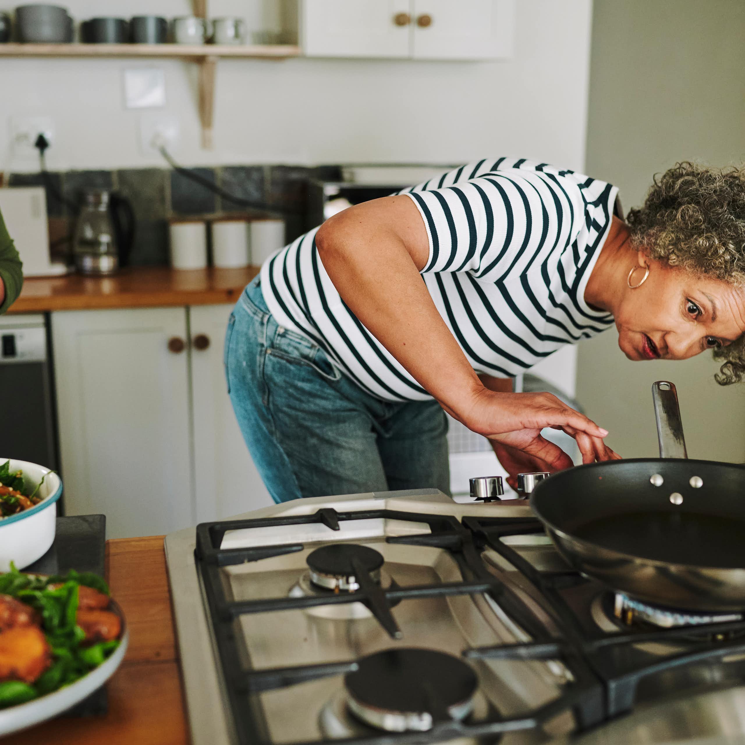 A woman with a black and white striped shirt looks under a pan on a stove top.