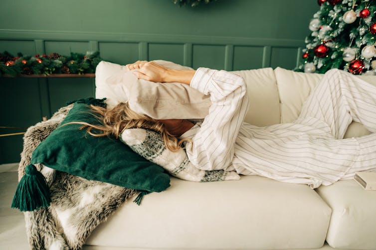 A stressed woman lying on a sofa, surrounded by Christmas decorations, hides her face in a pillow.