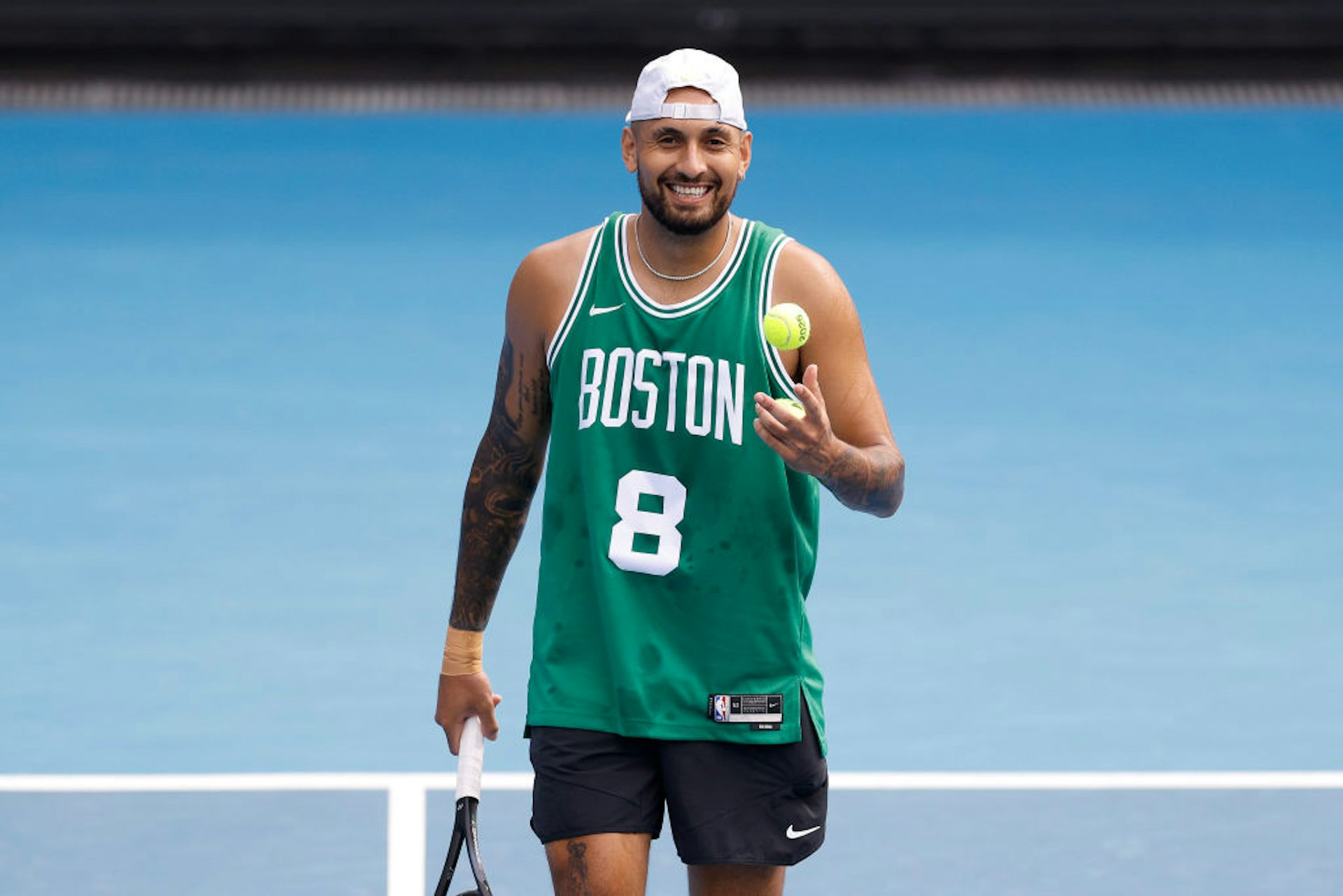Young man smiling while holding a tennis racket and a tennis ball, wearing a green basketball jersey and a backward white cap.