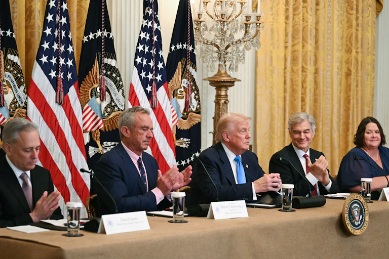 Trump seated on dais with two people on either side and American flags behind
