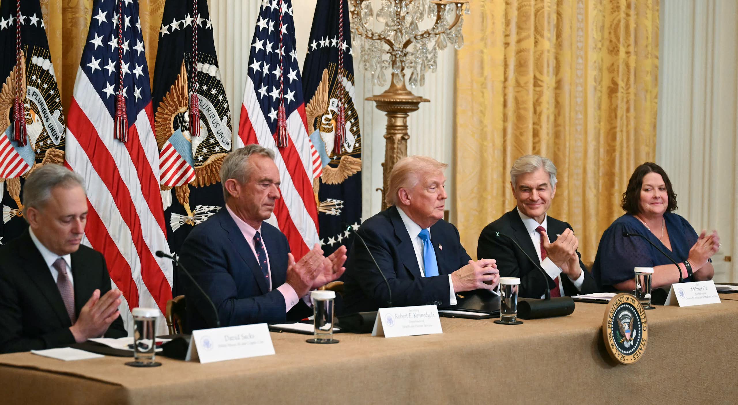 Trump seated on dais with two people on either side and American flags behind