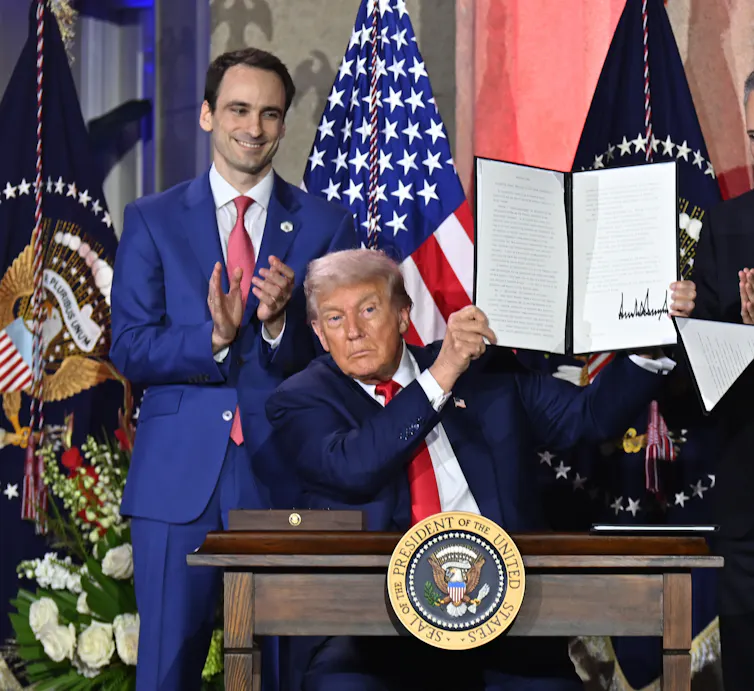 Michael Kratsios stands behind Trump, seated and holding up a signed document