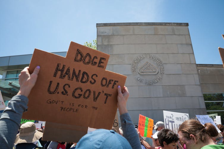 hands hold up cardboard sign 'DOGE HANDS OFF US GOVT' in front of NIH building