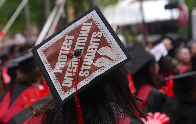 back of a graduating crowd with 'PROTECT INTERNATIONAL STUDENTS' on top of mortarboard