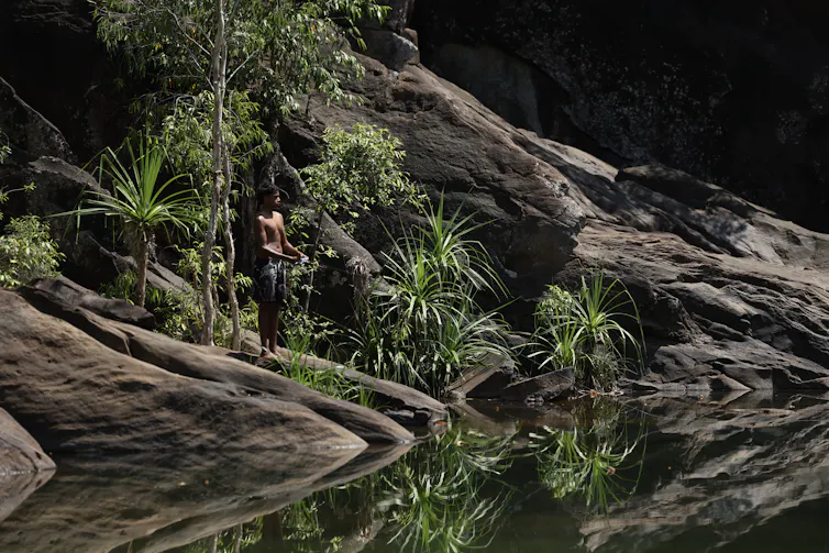 A young man holding a spear stands on the edge of a pool