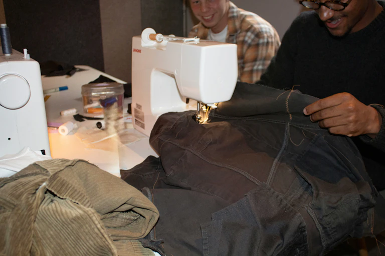 Two young men sit side by side in front of a table. The one in front is sewing a pair of jeans on a machine.