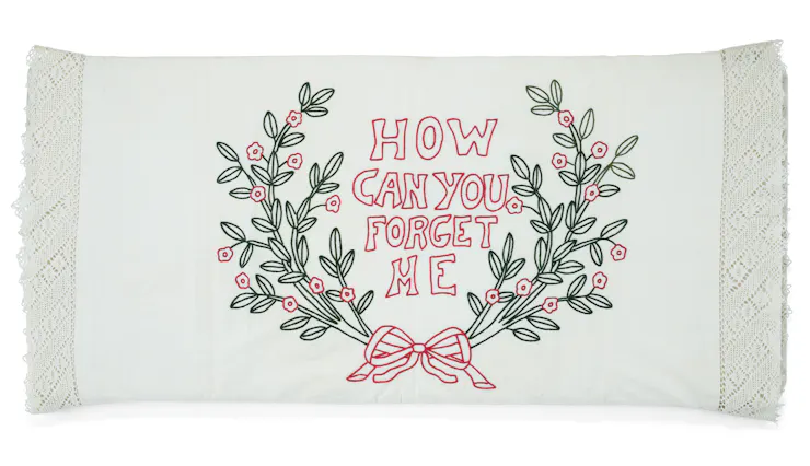 A white pillowcase with embroidery depicting a bow, green plants and the text 'How Can You Forget Me.'