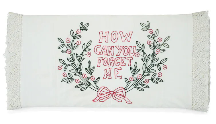 A white pillowcase with embroidery depicting a bow, green plants and the text 'How Can You Forget Me.'