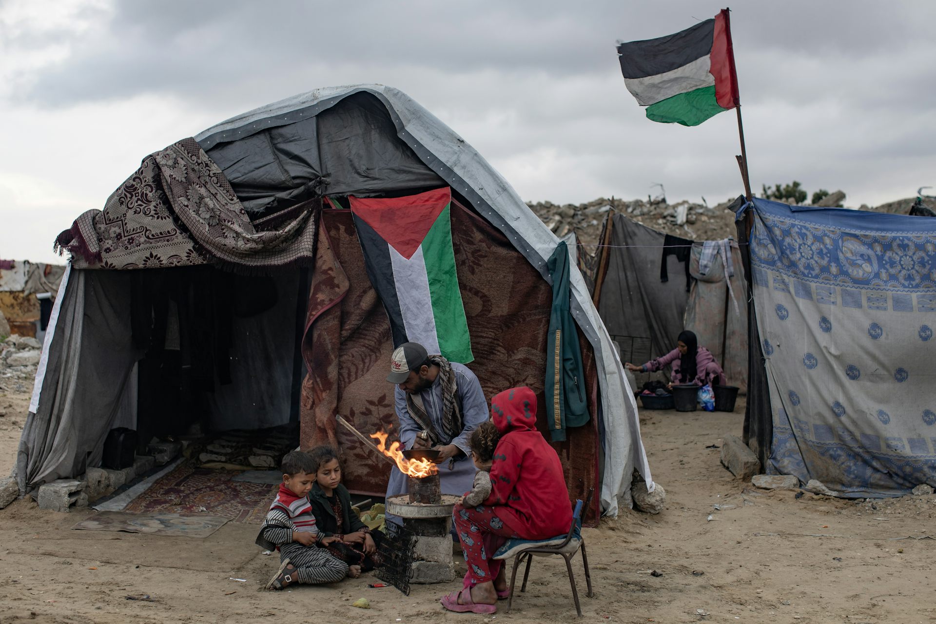Palestinians dressed in warm clothing outside a makeshift shelter with a Palestinian flag in the background.