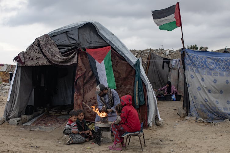 Dispatches from a 12 months of world battle and uncertainty 1 Palestinians dressed in warm clothing outside a makeshift shelter with a Palestinian flag in the background.