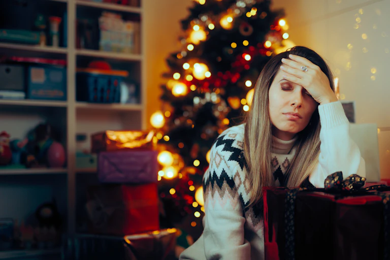 Stressed looking woman holds her head while sitting on the sofa with Christmas tree behind her