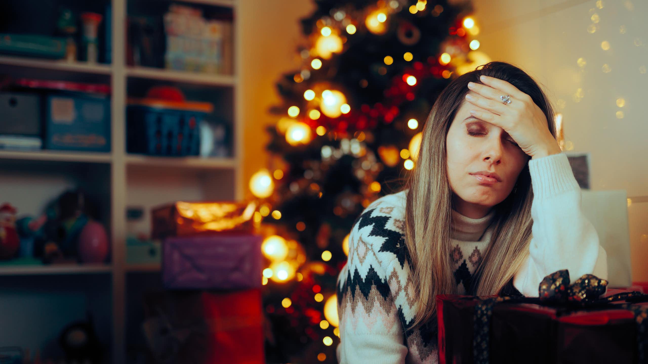 Stressed looking woman holds her head while sitting on the sofa with Christmas tree behind her