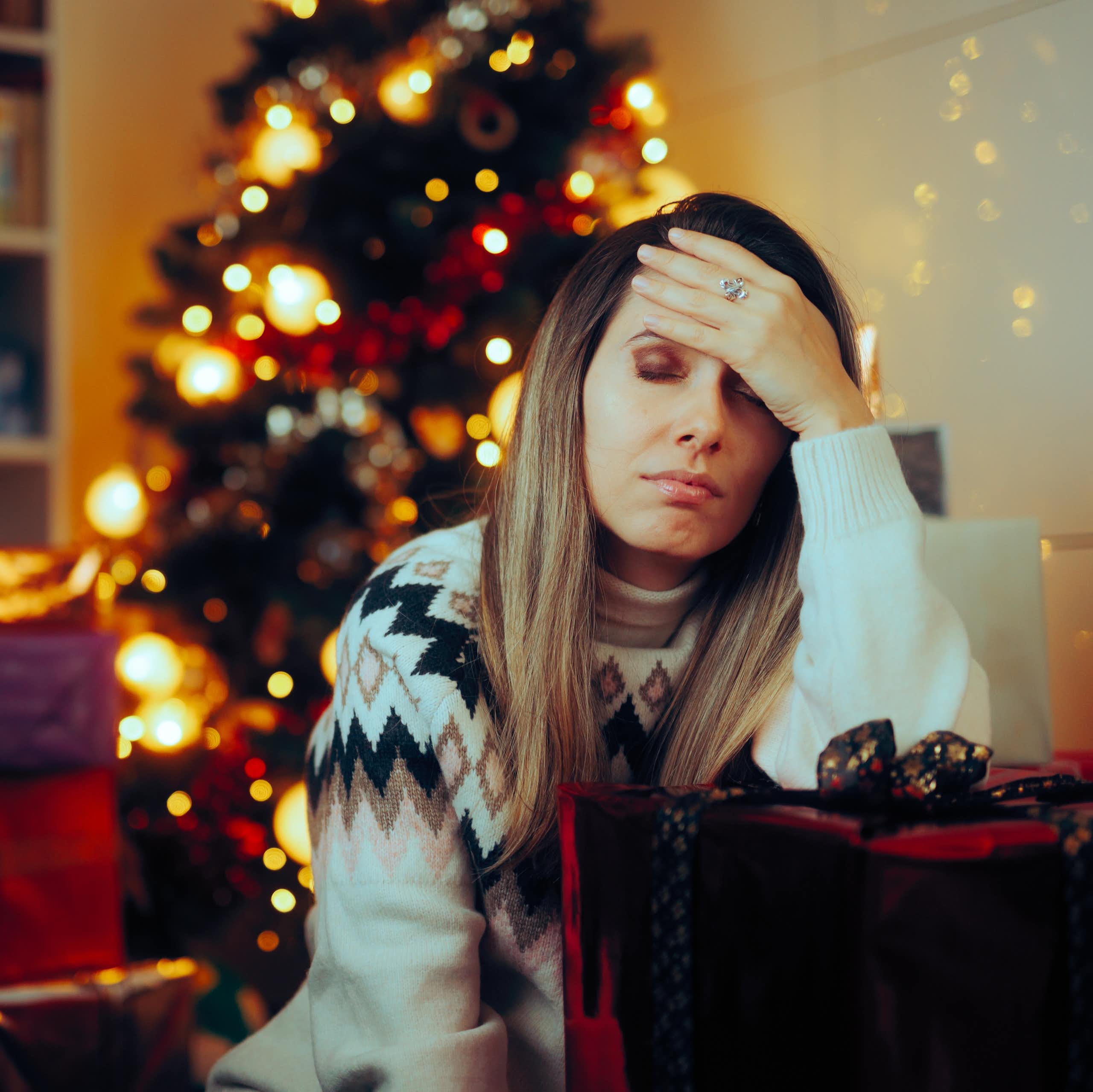 Stressed looking woman holds her head while sitting on the sofa with Christmas tree behind her