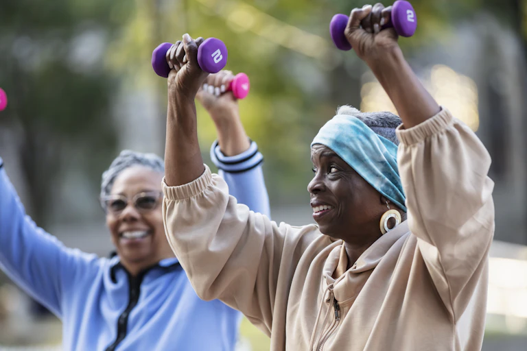 Two senior women smile while raising small pink and purple dumbbells in the air