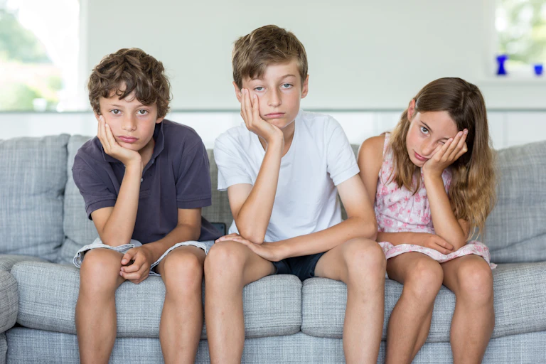 Three children sit on a couch together and all appear bored, with their faces resting in their hands.