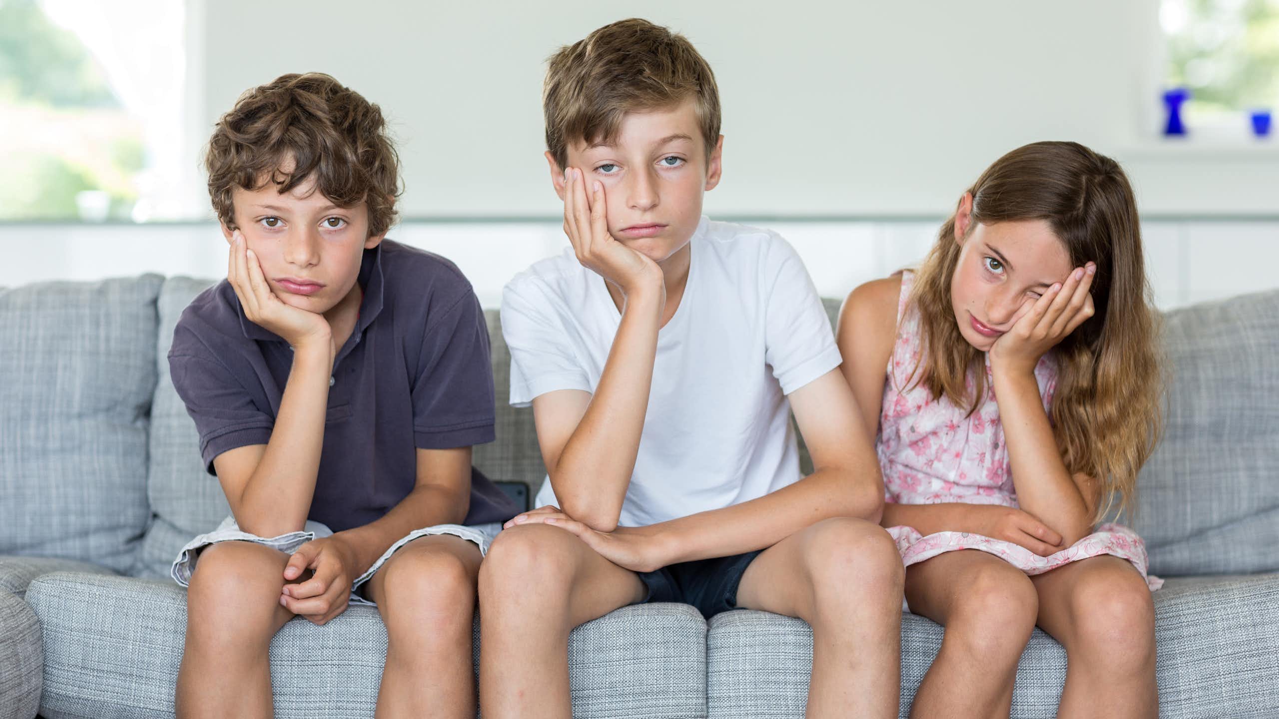 Three children sit on a couch together and all appear bored, with their faces resting in their hands.