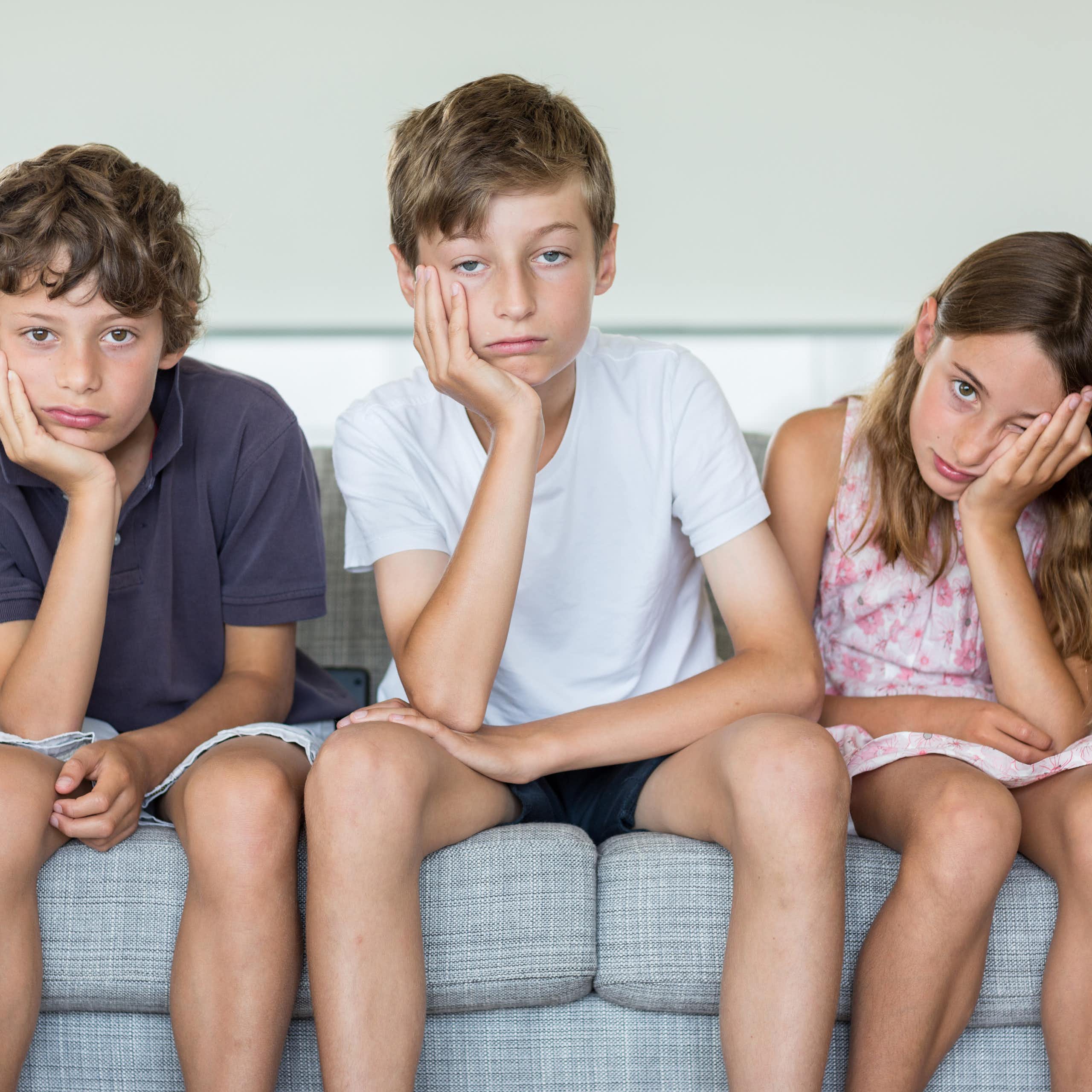 Three children sit on a couch together and all appear bored, with their faces resting in their hands.
