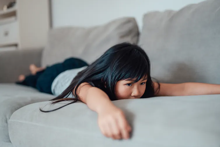 A young girl with dark hair lays on her stomach on a couch with her arms and legs splayed out.