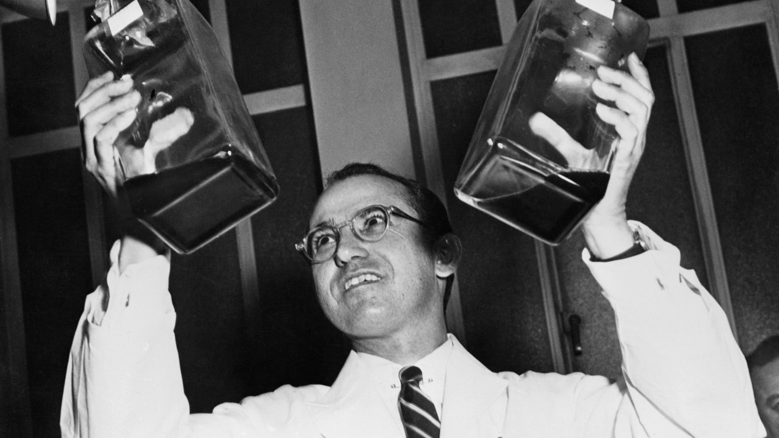Man in white lab coat holds up two large glass bottles containing a dark liquid