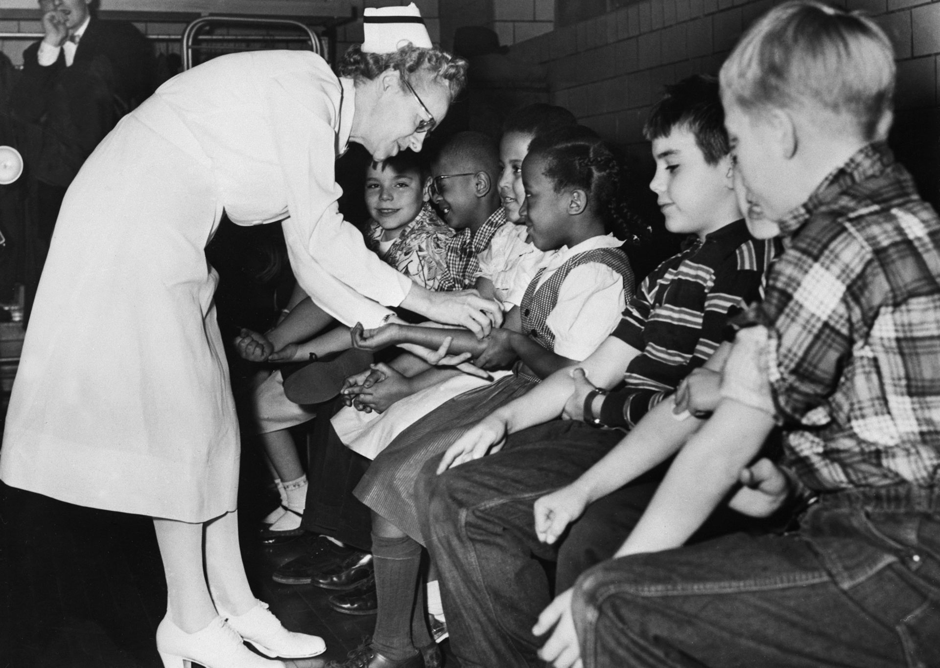 Black and white photo of nurse in white leaning over row of seated elementary school-aged children with sleeves rolled up