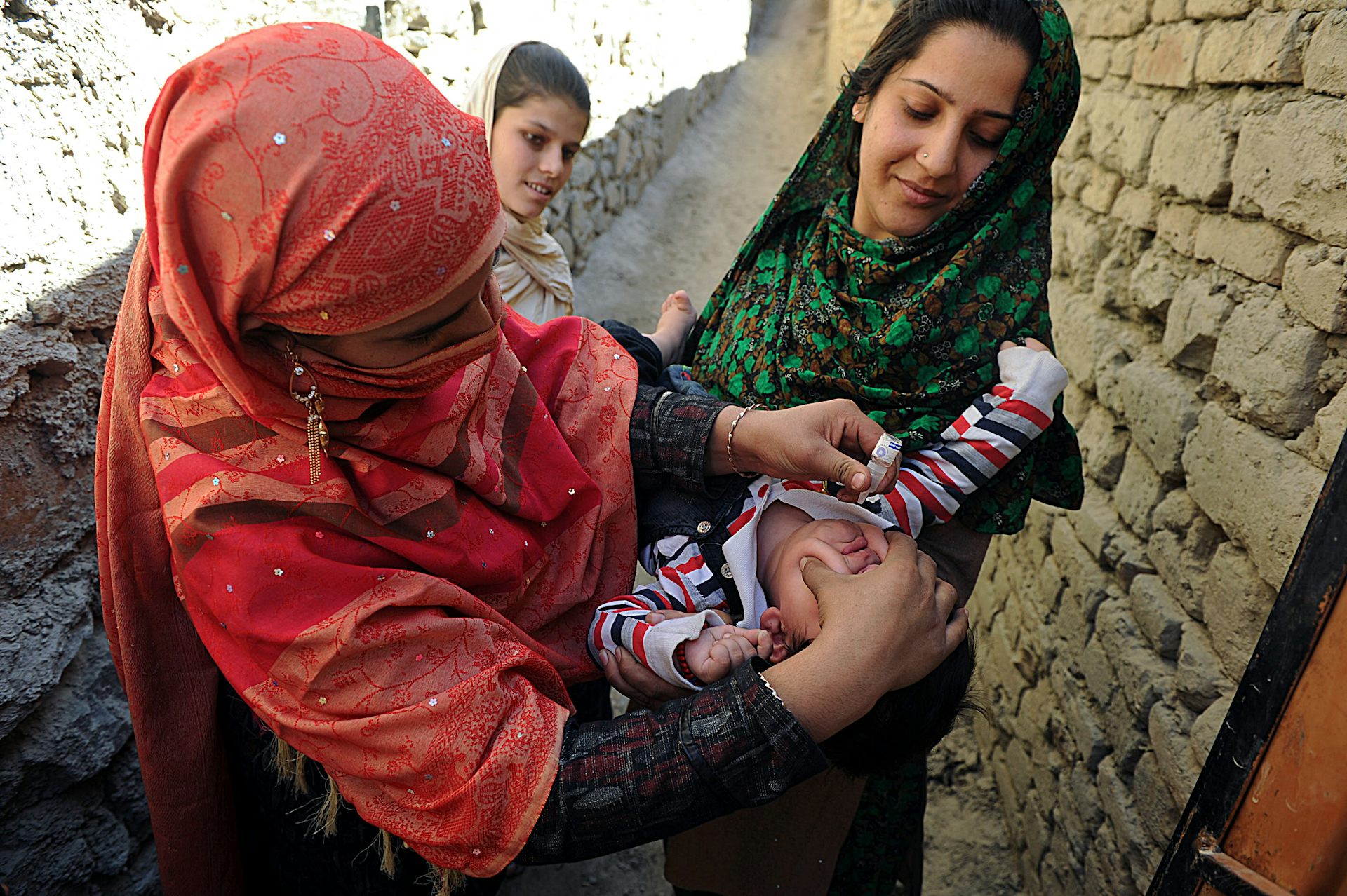 Woman in traditional Afghan dress tries to squeeze medical drops into mouth of a baby held in another woman's arms