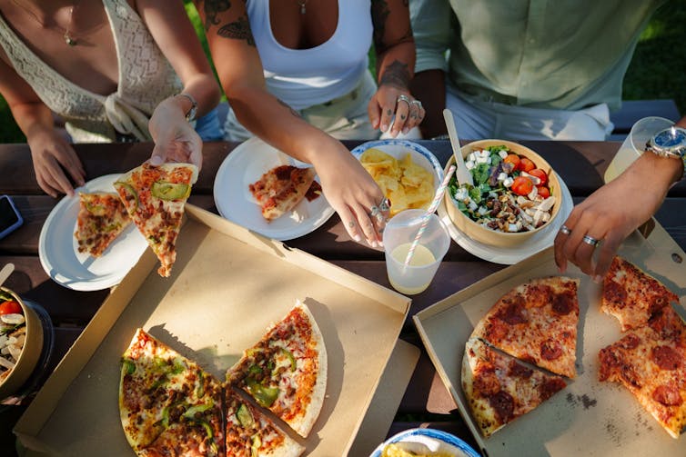 Three people are filling their plates with pizza, salad and chips
