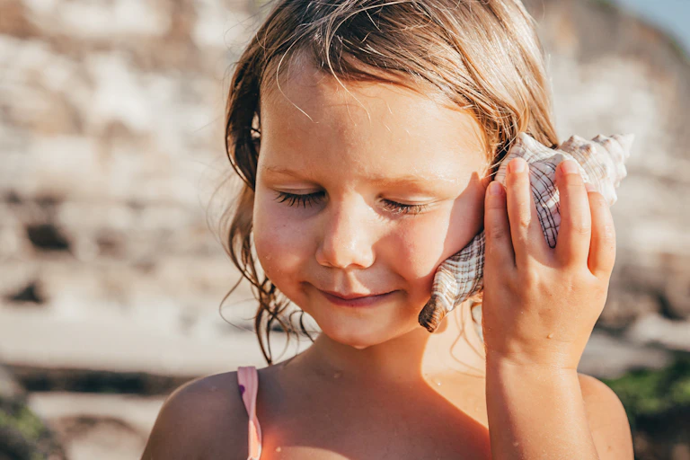 young girl at the beach holds a seashell up to her ear