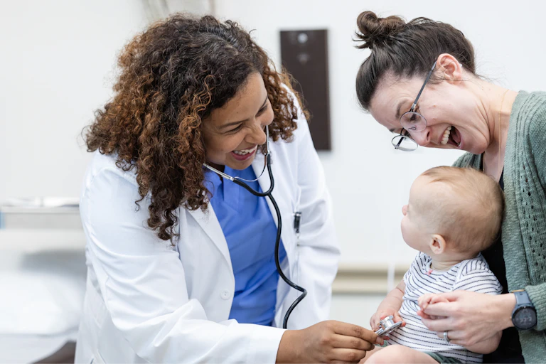 Young mother smiles and tries to distract her baby while the doctor is examining the baby.