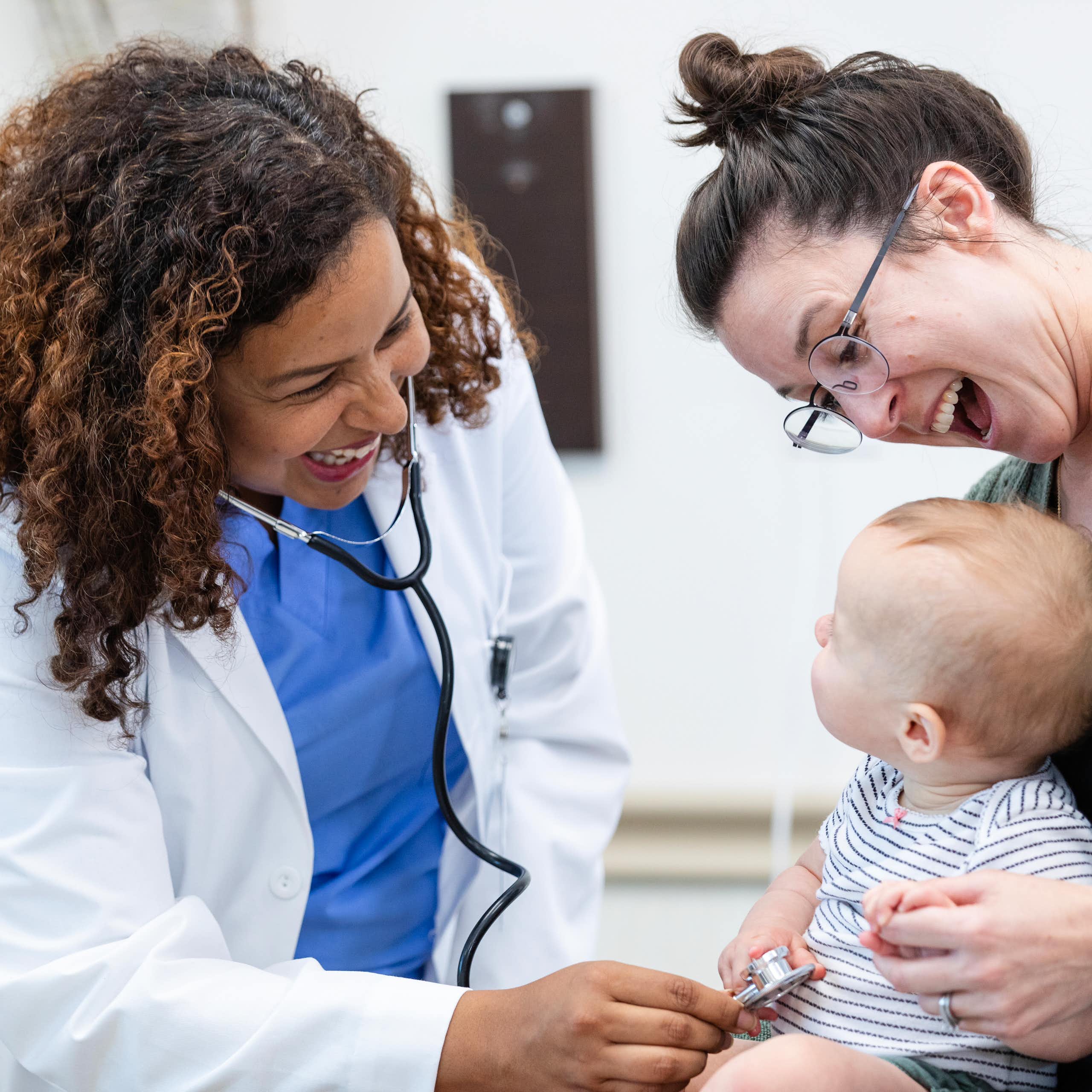 Young mother smiles and tries to distract her baby while the doctor is examining the baby.