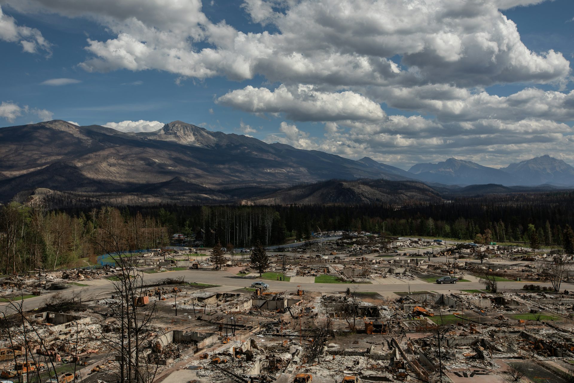 an aerial photo of destroyed buildings and rubble, mountains under a blue sky with white clouds in the background.