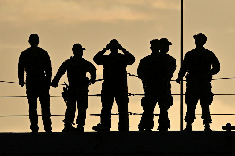 Five military personnel in silhouette, standing on the deck of a ship.