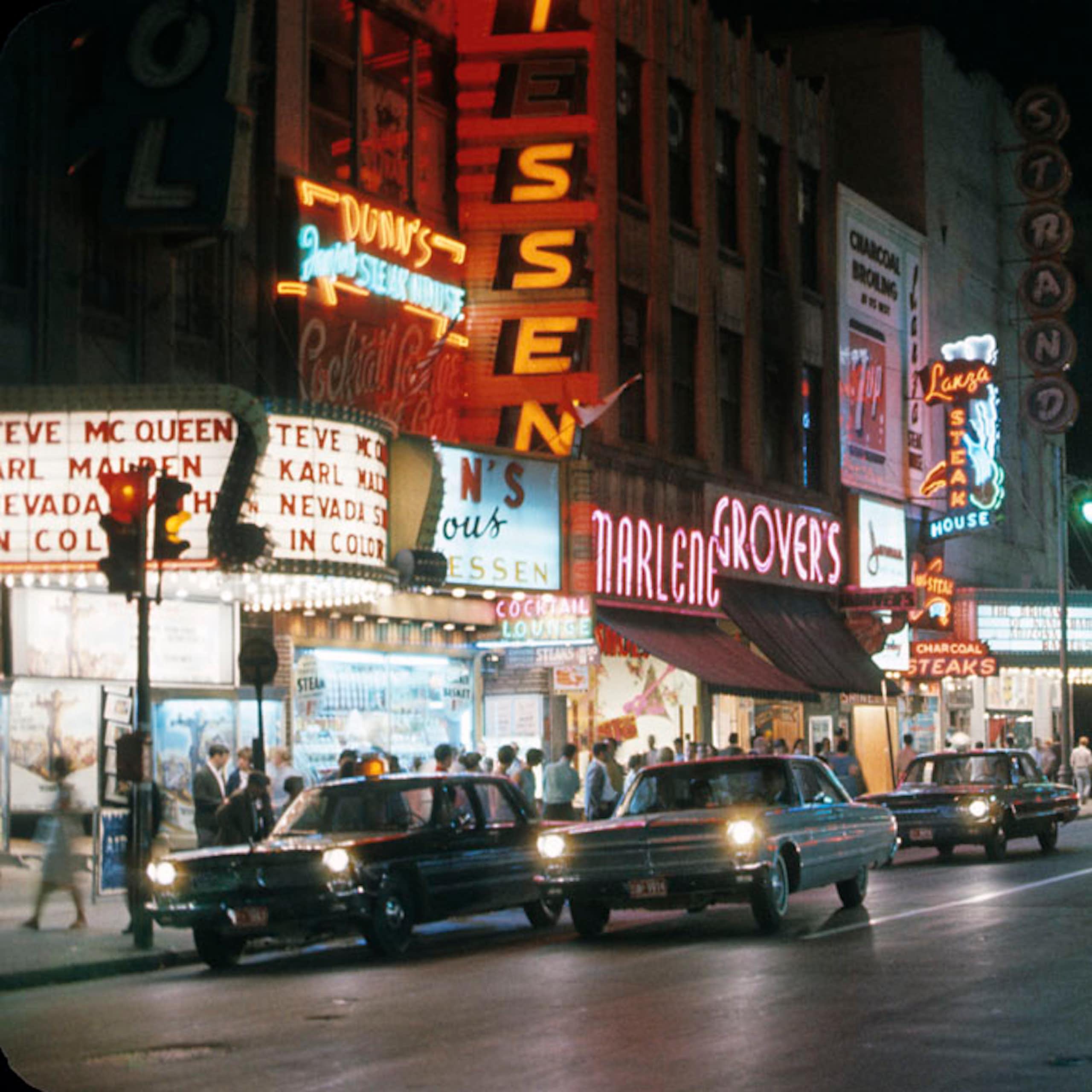 Lumières néon dans la nuit, avec des voitures circulant dans le centre-ville et des passants marchant sur les trottoirs.