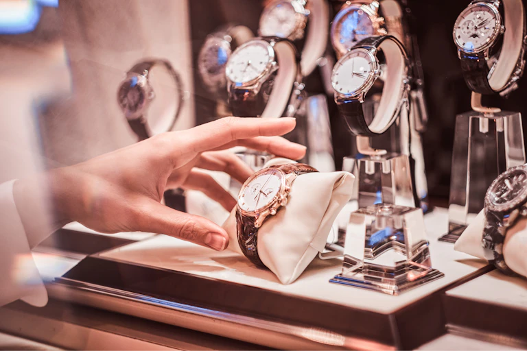 luxury watches on display in a shop window with a hand reaching out to take one.